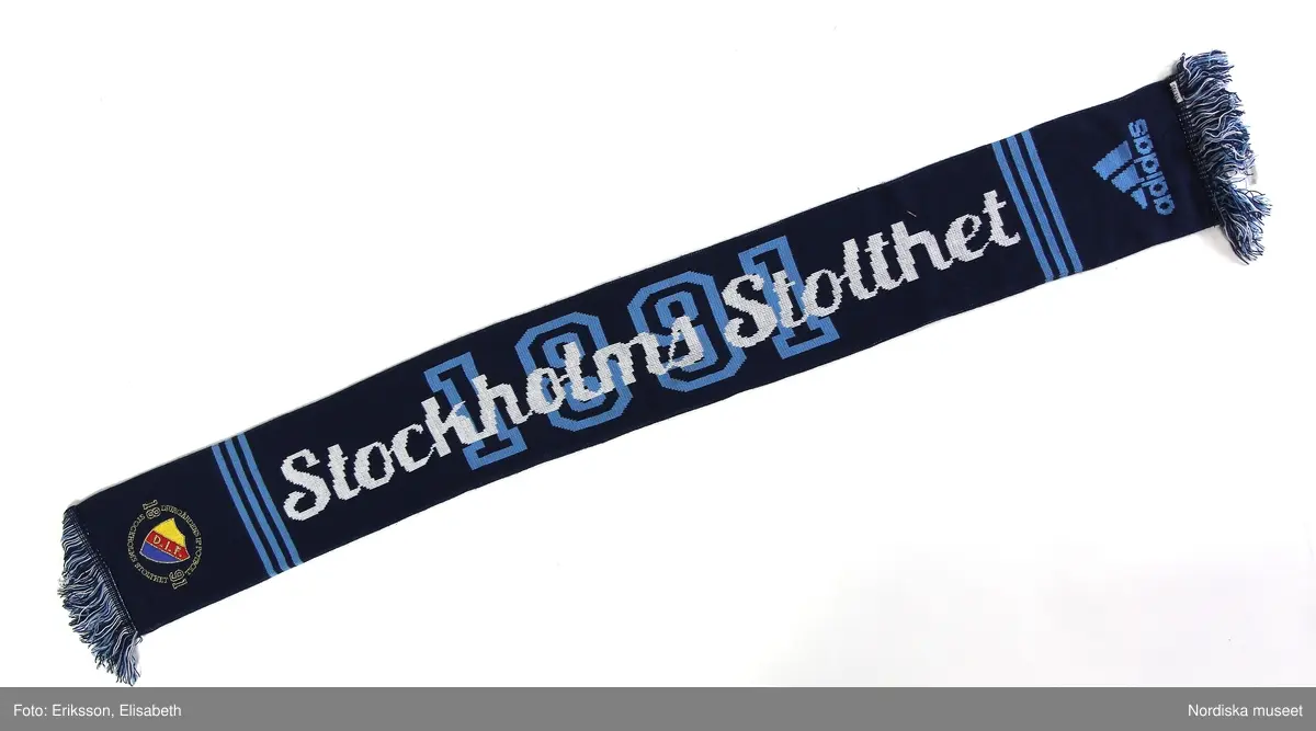 Föreningshalsduk från Djurgårdens idrottsförenings (DIF) med färgerna ljus- och mörkblått. Med fransar.
Några med Djurgårdens emblem i gult-rött-blått.
Längden mellan ca 120-160 cm.
Bredd ca 15-20 cm.

2019-02-21 Cecilia Hammarlund-Larsson/Lena Kättström Höök