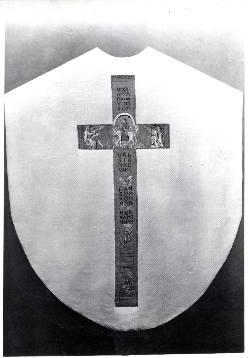 Foto (svart/vitt) av en ljus mässhake (baksidan) i brokad (?), med mörkare, broderat korsparti, prytt bl a med Jesus på en åsna och text; "Hosianna Davids Son, Välsignad Vare Han Som Kommer i Herrans Namn".

Inskrivet i huvudbok 1983.