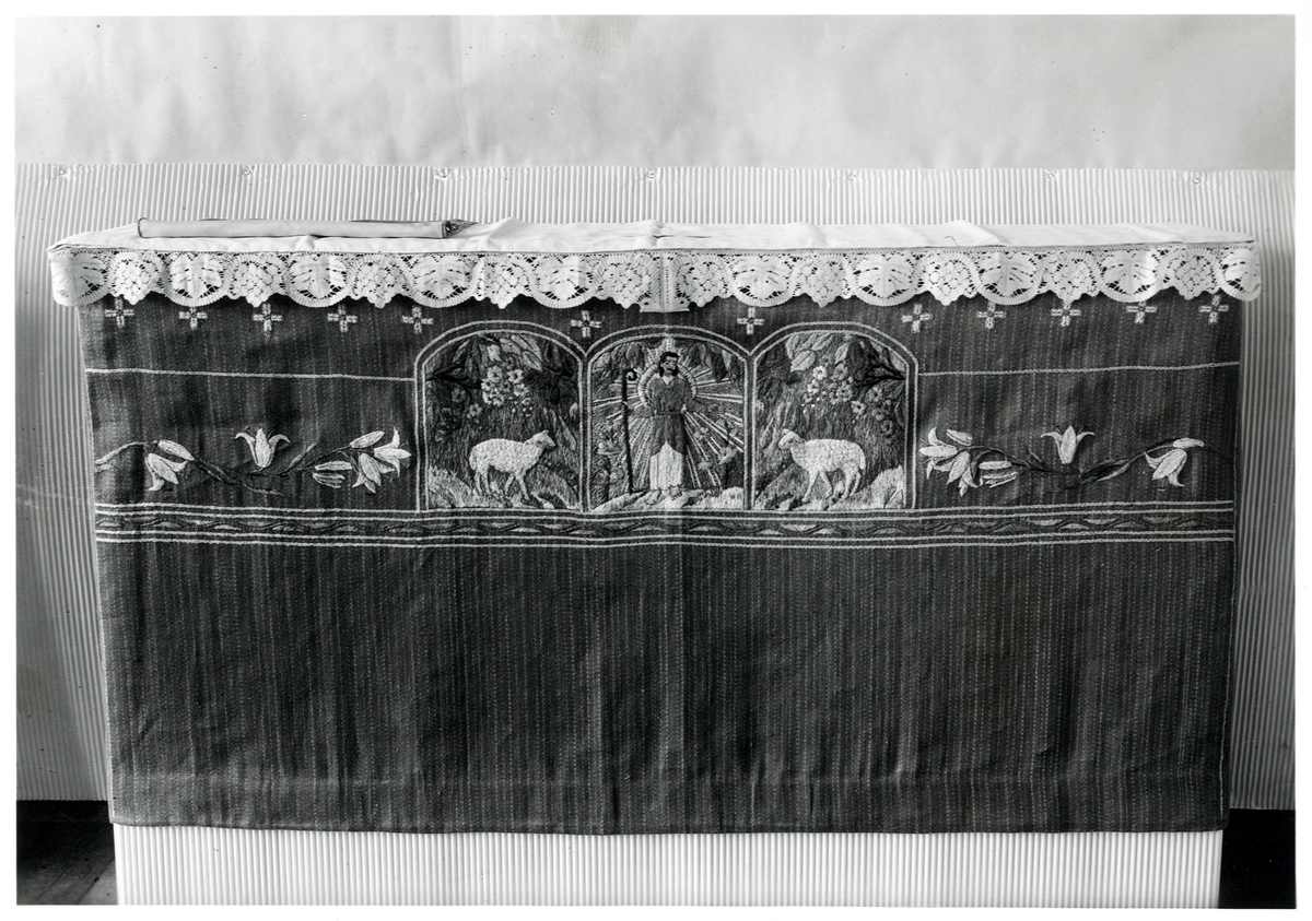 Foto (svart/vitt) av ett mörkt antependium med broderier i form av en herde med lamm och vita liljekvistar.

Inskrivet i huvudbok 1983.