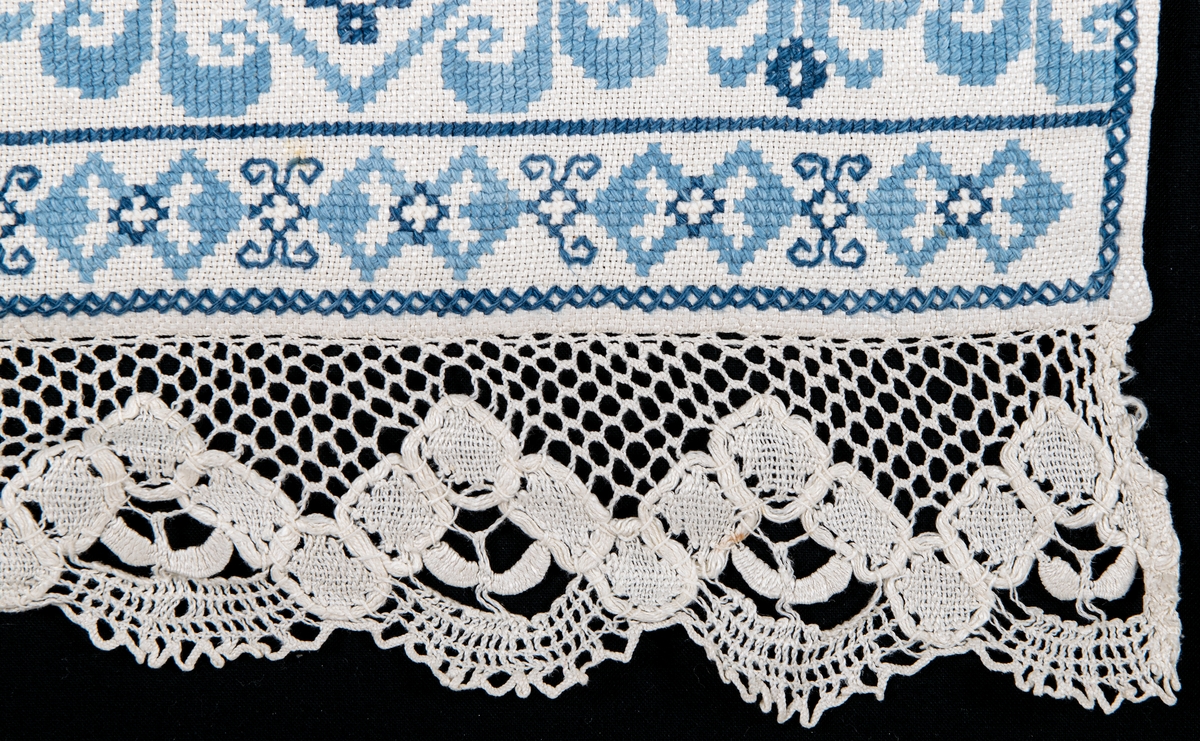 Serveringsbordsduk i vitt linne, broderad med blått korssömsbroderi i två nyanser. Framkanten i knypplad spets.