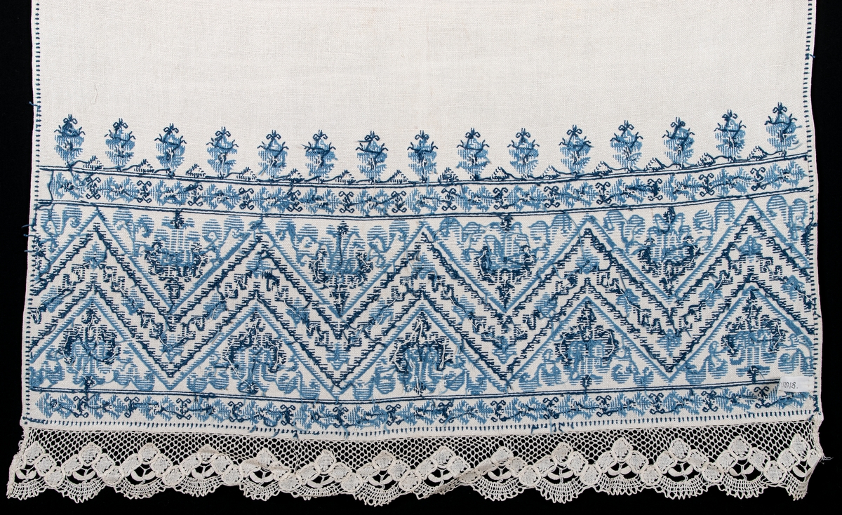 Serveringsbordsduk i vitt linne, broderad med blått korssömsbroderi i två nyanser. Framkanten i knypplad spets.