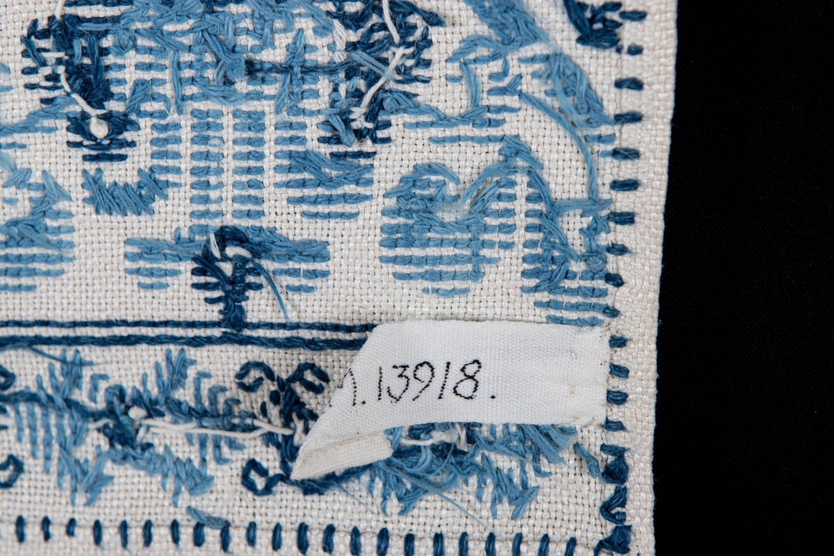 Serveringsbordsduk i vitt linne, broderad med blått korssömsbroderi i två nyanser. Framkanten i knypplad spets.