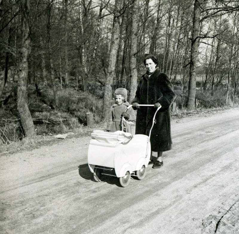 Eivor Bjerrhede (1921 - 2015) är på promenad med dottern Annika (född 1948) och lille Staffan (född 1953) liggandes i barnvagnen. De går på en väg vid Tulebosjön år 1953.