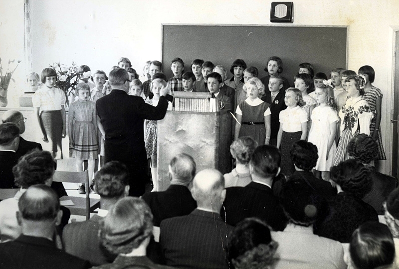 Invigning av Kålleredskolan (senare Brattåsskolan) 1954 eller 1955.
Folkskollärare och kantor Gunnar Alander leder en kör av elever. Föräldrar och politiker bland publiken.