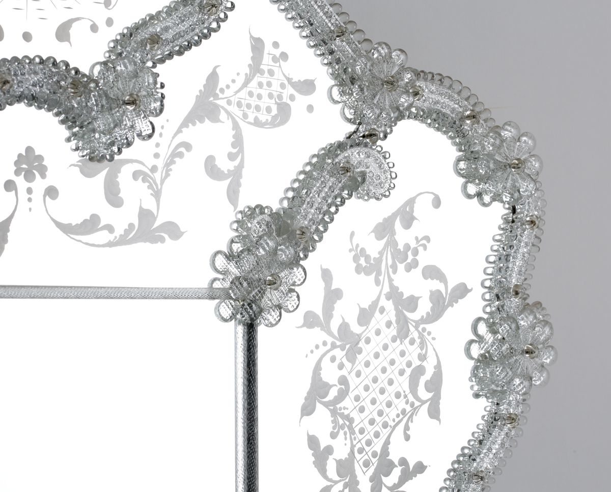 Spegel i venetiansk stil. Ram av skulpterade partier och blommor. Stiliserade graverade växtmotiv runt om, men vid krönet ett kärlekspar i 1700-talsdress.