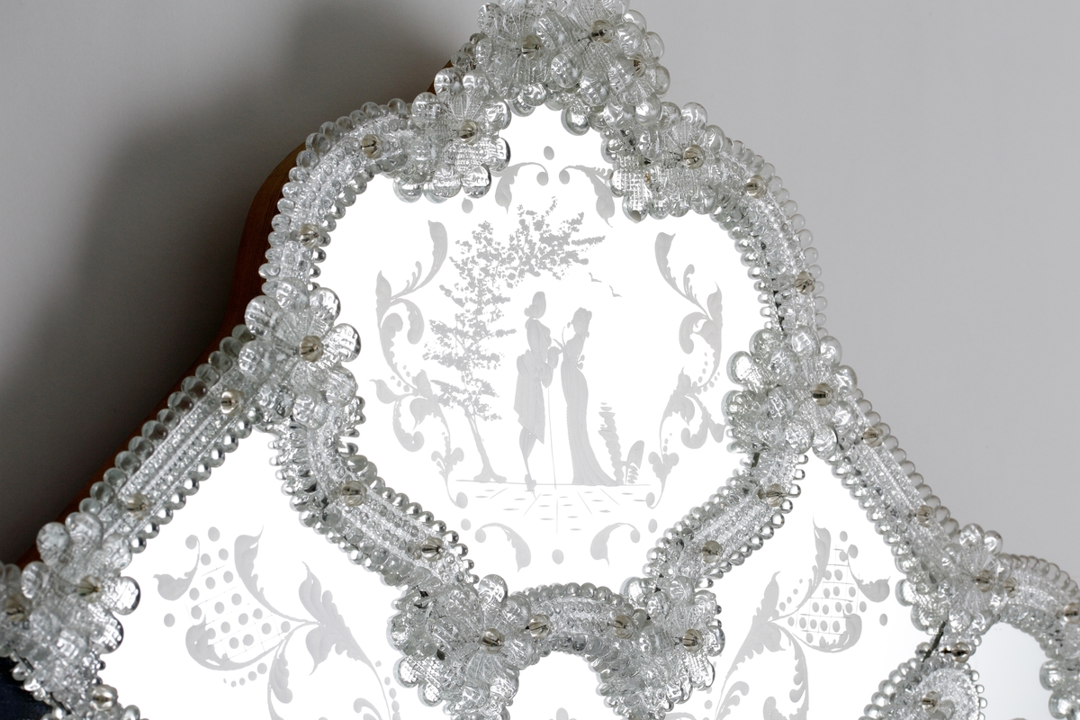 Spegel i venetiansk stil. Ram av skulpterade partier och blommor. Stiliserade graverade växtmotiv runt om, men vid krönet ett kärlekspar i 1700-talsdress.