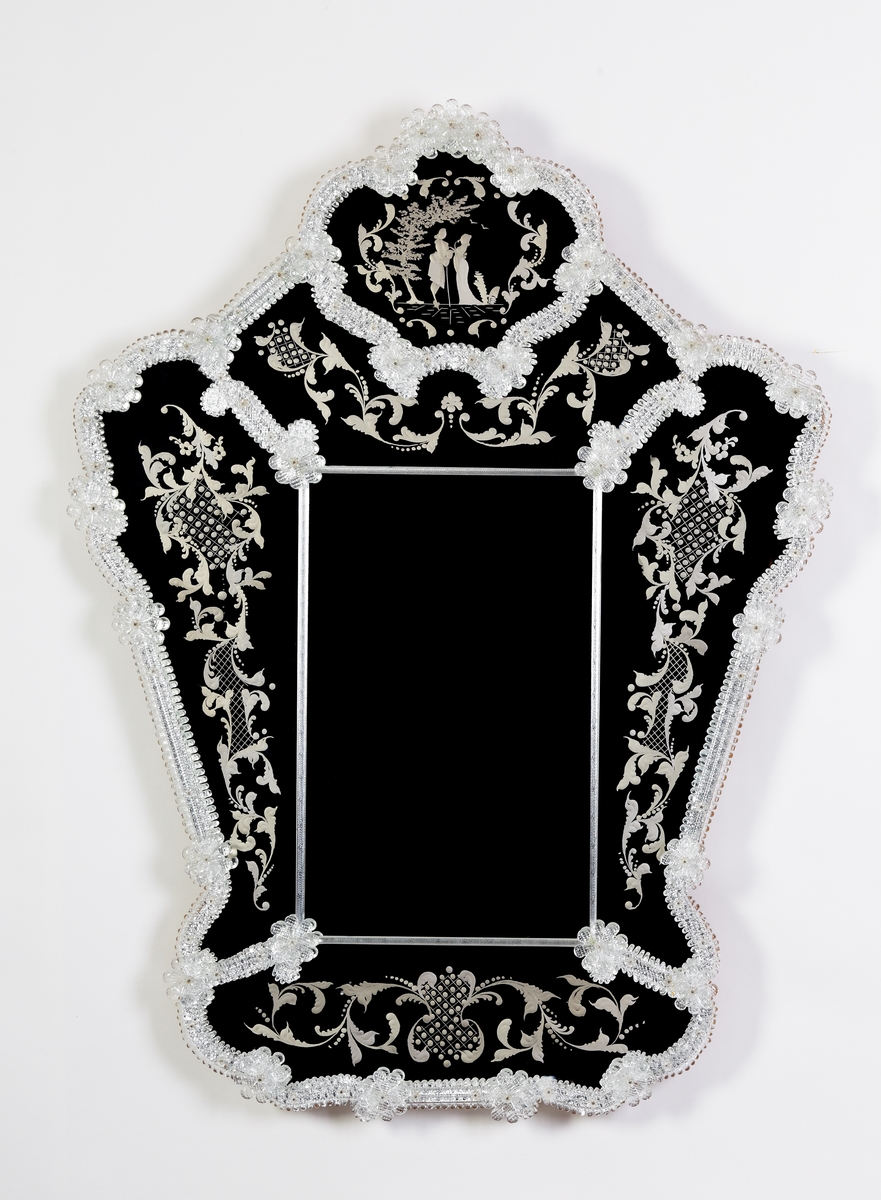 Spegel i venetiansk stil. Ram av skulpterade partier och blommor. Stiliserade graverade växtmotiv runt om, men vid krönet ett kärlekspar i 1700-talsdress.