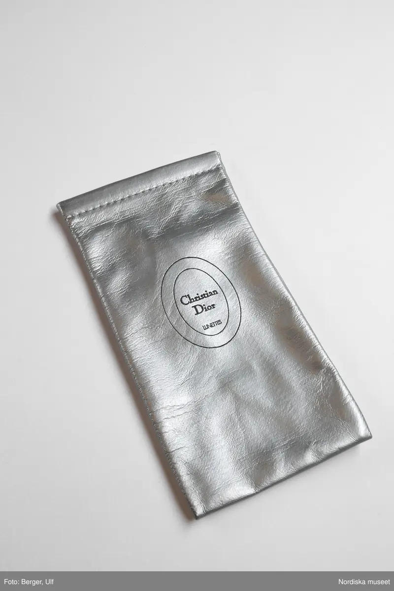 Ett glasögonfodral i silverfärgad plast med bomullsväv som foder. Öppning vid övrekanten med en mjuk metallband för stängning. "Christian Dior LUNETTES" stämpladt i skinnet inom två ovaler placerades på mitten av framsidan. 
/Petrine Knight, 2014-12-09
