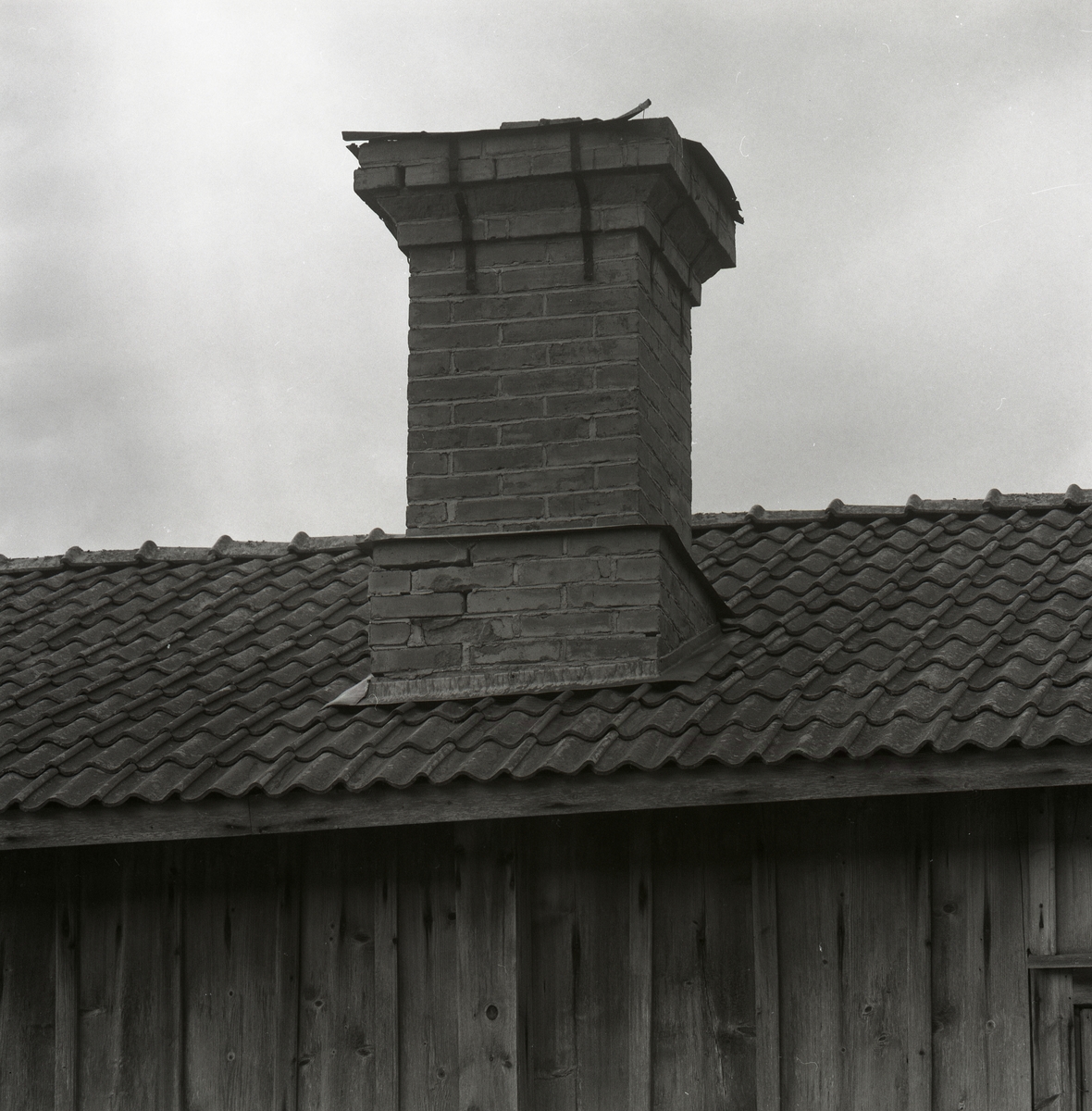 En träbyggnad med tegeltak och murad skorsten och himlen i bakgrunden. Utegårn 24 augusti 1990.