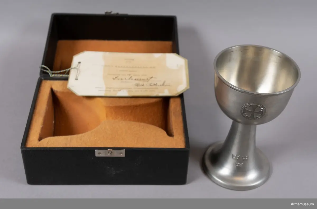 Modellexemplar av nattvardskalk m/1940, större modell i tenn. Märkt "N Redeby" inuti foten. Förvaras tillsammmans med paten i svart fodral försett med sigill samt etikett signerad av byråchefen för Kungliga arméförvaltningens intendenturdepartement den 15 april 1940.