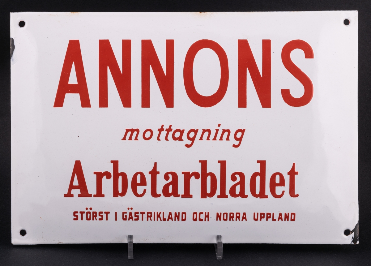 Emaljerad plåtskylt. Rektangulär, något kupad med skruvhål i hörnen. Vit med röd text: Annonsmottagning, Arbetarbladet. Störst i Gästrikland och norra Uppland.