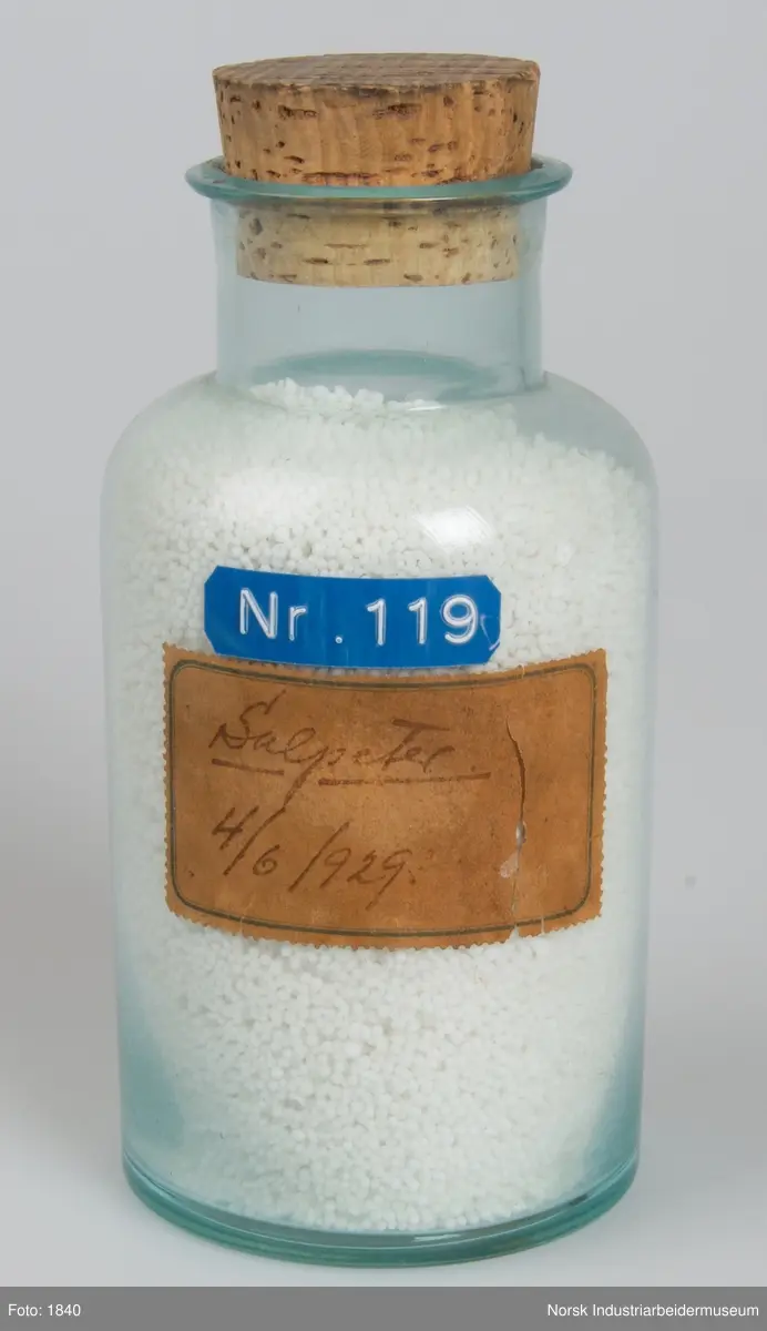 Laboratorieglass med prøve av den første produksjonen av kalksalpeter.
Cylindrisk laboratorieglass med korkpropp. Inneholder salpeter i form av små hvite perler.
Glasset er forsynt med selvklebende blått plastmerke (Dymo). NR. 119.