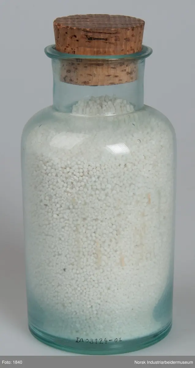 Laboratorieglass med prøve av den første produksjonen av kalksalpeter.
Cylindrisk laboratorieglass med korkpropp. Inneholder salpeter i form av små hvite perler.
Glasset er forsynt med selvklebende blått plastmerke (Dymo). NR. 119.