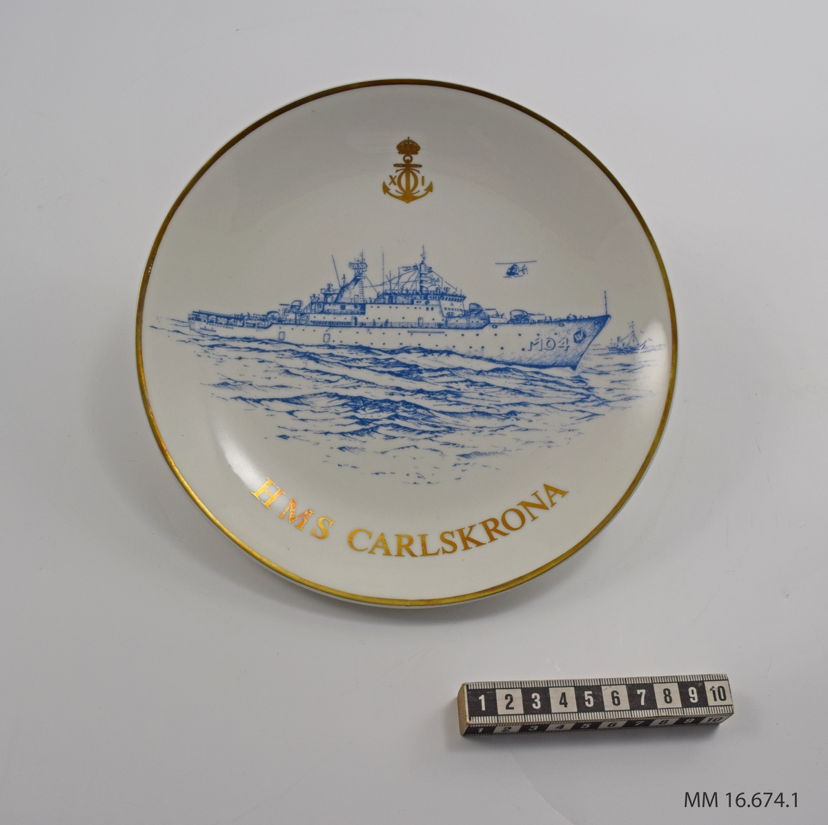 Minnestallrik av vitt porslin. Dekoration: i mitten ses HMS Carlskrona under gång, en lågt flygande helikopter, i bakgrunden ett mindre fartyg. Motivet - utförd i blått. Över motivet: Karlskronas emblem i guld. Runt tallrikens kant går ett guldband. Baksidan: Flottans mäns emblem och text samt Gustavsbergs emblem. Allt i guld.