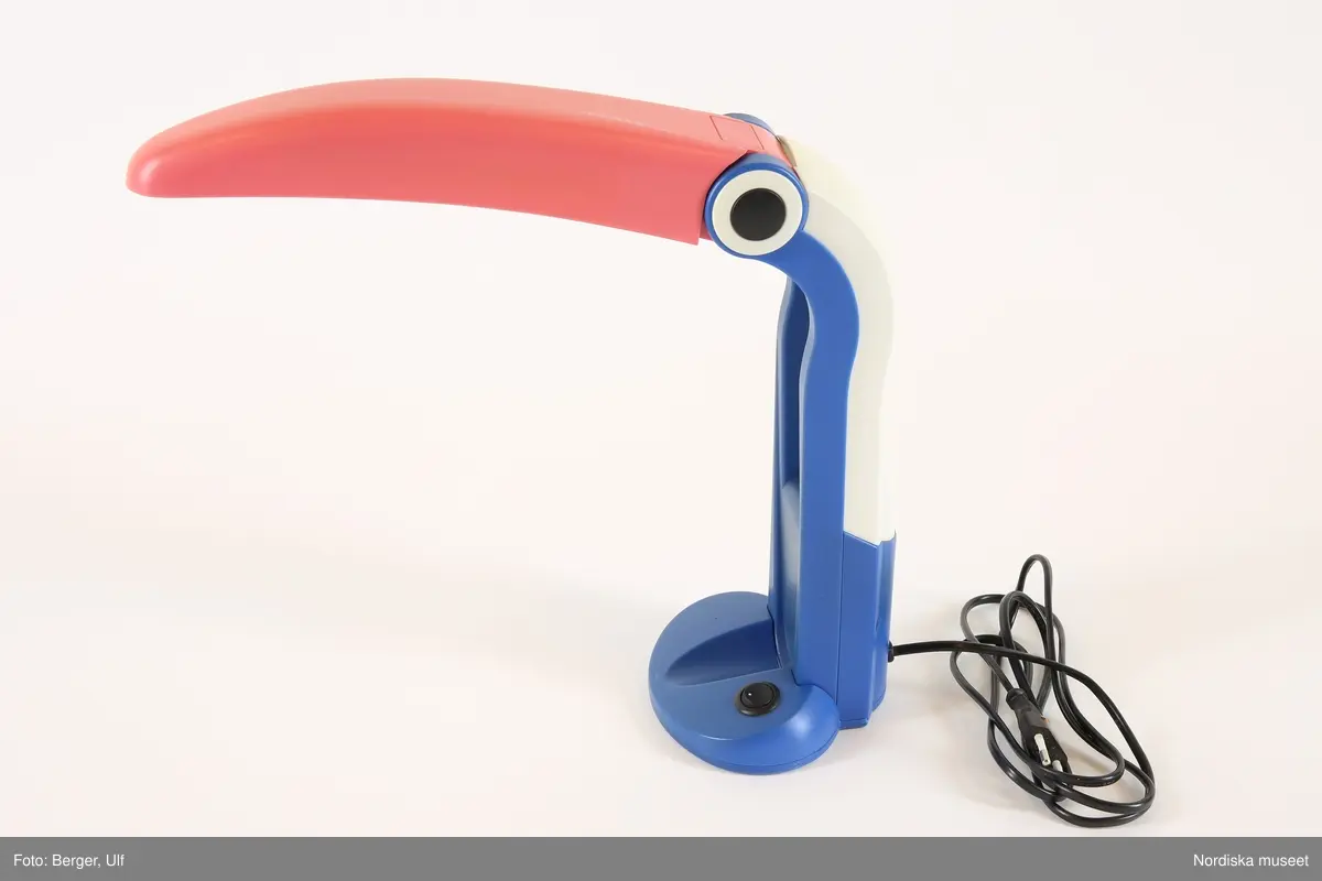 Skrivbordslampa av plast med formen av en tukan/pelikan. Rund fot, vertikalt plastskaft i blått och vitt, upptill med mjuk böj avslutad i cirkelrunt gångjärn med formen av ett stort runt öga i svart med vit ytterkontur. På andra sidan "ögat" rödfärgad lång näbb med reflektor och lysrörslampa monterade undertill. Lampan kan vikas ihop genom att näbben viks rakt ned och då blir parallell med lampans skaft.
/Maria Maxén 2017-06-29