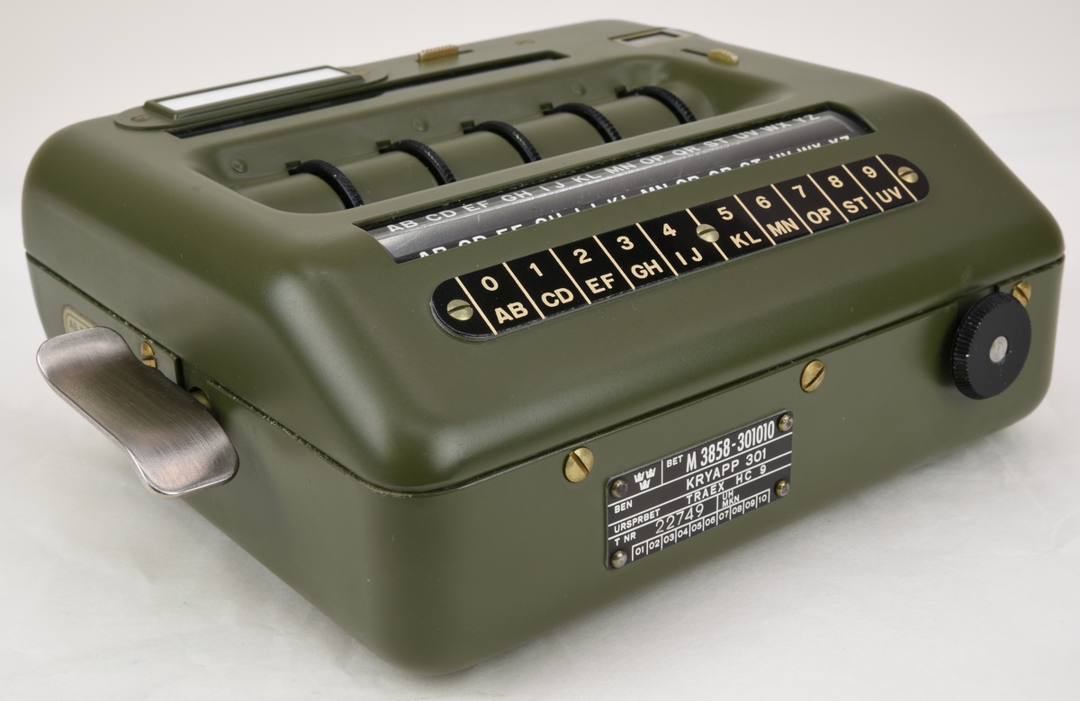 Krypteringsapparat (:1) med hölje av grönmålat aluminium i rundade former. Översidans framkant är lätt fasad och har ett avlångt fönster med alfabetet i över- och nederkant. Fönstret är till för att visa den alfabetsrulle som gav lösningen på kryptot. Under fönstret finns en svart skylt med vit text där siffrors bokstavskoder visas. På ovansidans mitt finns fem smala chifferhjul av svart bakelit i en försänkning. Hjulen är till för att ställa in chiffrets bokstäver som syns i ett fönster längst upp på apparatens ovansida. Till höger om chifferfönstret finns ett räkneverk med fyra siffror.

På apparatens framkant finns en skylt med M-nummer, serienummer och liknande och till höger ett vred av svart bakelit för att mata fram alfabetsrullen. På apparatens vänstra sida finns ett frammatningsvred av metall samt tillverkarens logotyp. På den högra sidan finns en lucka för alfabetsrullen och ett vred av bakelit för att ställa in chifferhjulaxeln. Baksidan har en större lucka för hålkortsplatsen och undersidan har små gummifötter samt en dekal med information om garantitid.

Till apparaten hör också en transportväska (:2) av formgjuten och genomfärgad gummi. Väskan är grön och har en vävd bärrem fäst med mässingsskruv. Locket är fäst med ett avlångt, nitat gångjärn och har ett låsspänne i mässing. På locket finns tre kronor och apparatens M-nummer i relief och på väskans undersida finns väskans M-nummer. Insidan av väskan är formad efter kryptoapparaten för att ge så mycket skydd som möjligt.

M-nummer: M 3858-301010
Tillverkningsnummer: 22749

Väskans M-nummer: M 7038-045010