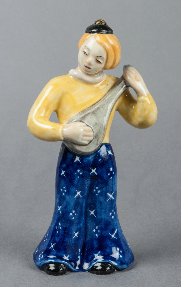 Figurin Zahra, kvinna spelande mandolin, glaserad i främst gult och blått. Tillverkad av Gefle Porslinsfabrik, formgiven av Märta Willborg 1937.