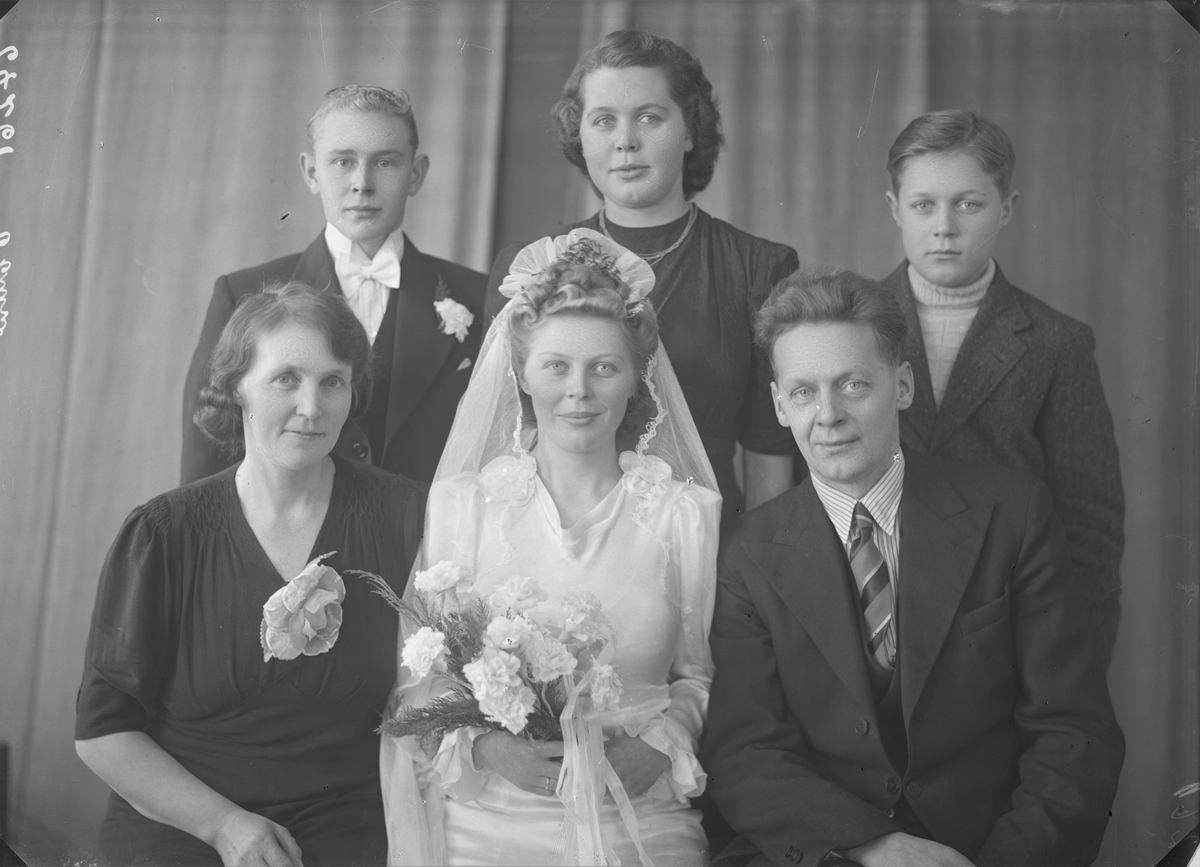 Gruppebilde. Familiegruppe på seks. Brudebilde. Ung kvinne i lang hvit brudekjole med slør som holder en blomsterbukett i hendene omkranset av  fem familemedlemmer. Mor og far ved siden og tre yngere søsken stående bak. Alle pyntet til fest. Bestilt av Morten Nilsen. Smørsund.