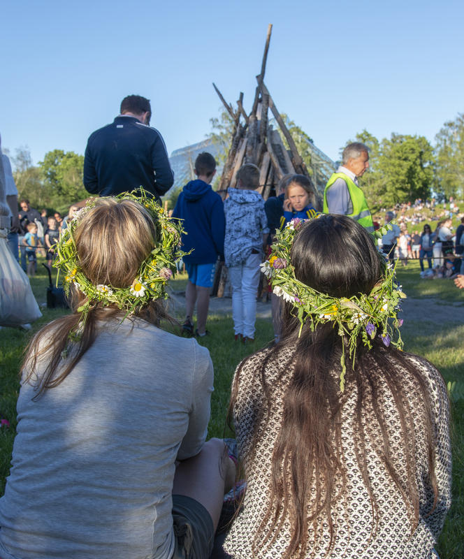 To jenter med langt hår og blomsterkrans på hodet sitter med ryggen mot fotografen og ser mot bålet som snart skal tennes.