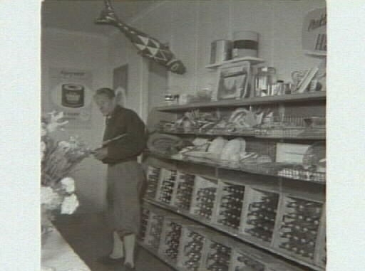 Affärsinteriör med staplade drickabackar i trä och en man i "äppelknyckarbyxor". "Kooperativa öppnar ny affär på Getterön". Artikel i samband med bilden publicerad i Varbergs Tidning 1955-06-15.