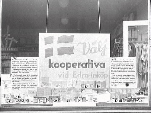Skyltfönster till Kooperativas butik i Varberg. "Välj kooperativa vid Edra inköp" står det på en stor skylt i fönstret. I bakgrunden syns en stång med kläder.