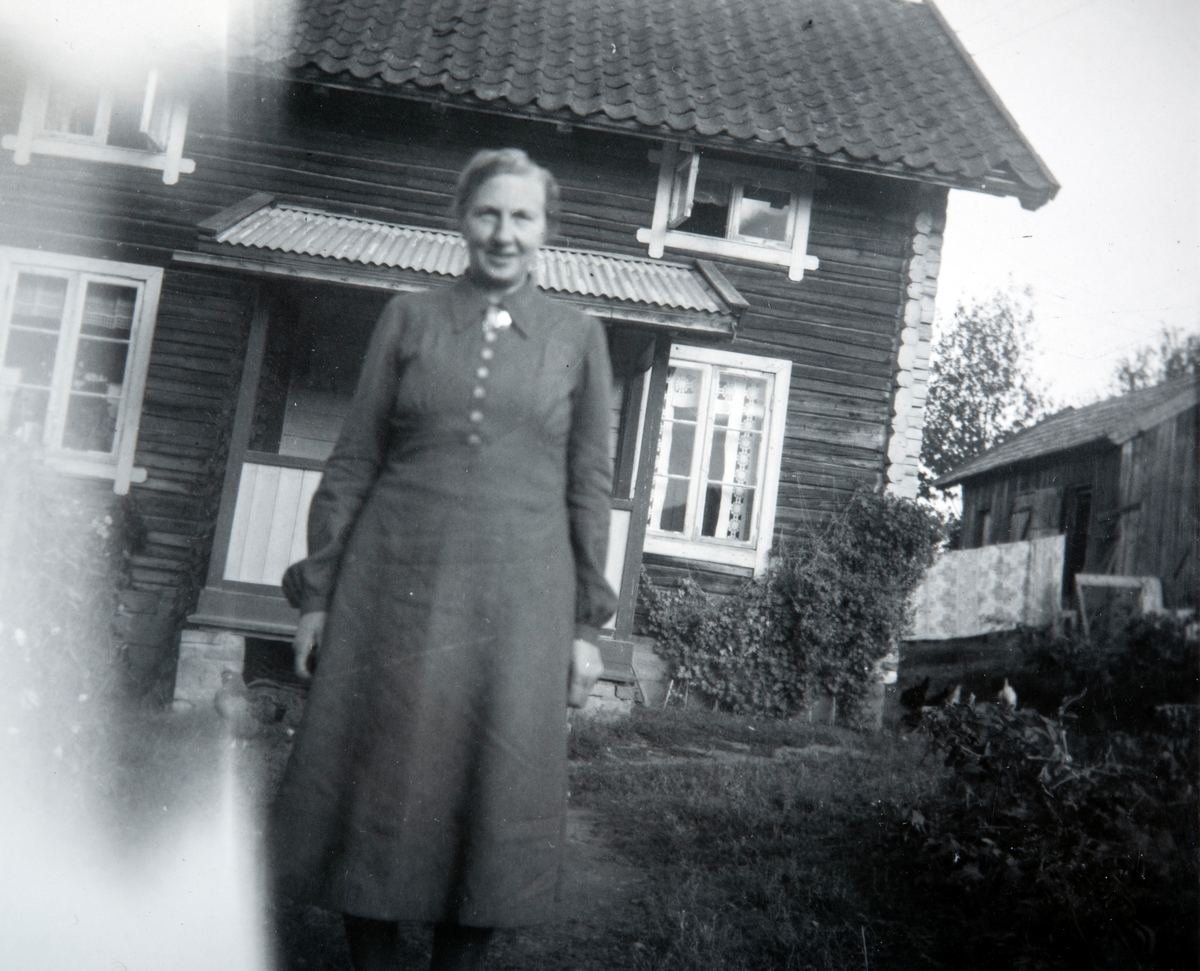 Ukjent dame på Kjøs, 81/1. Tømmerstue i bakgrunn.