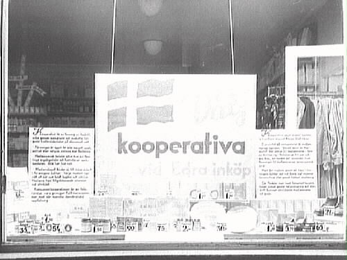 Skyltfönster till Kooperativa AB, butik i Varberg. "Välj kooperativa vid Edra inköp" står det på en stor skylt i fönstret. I bakgrunden syns en stång med kläder.