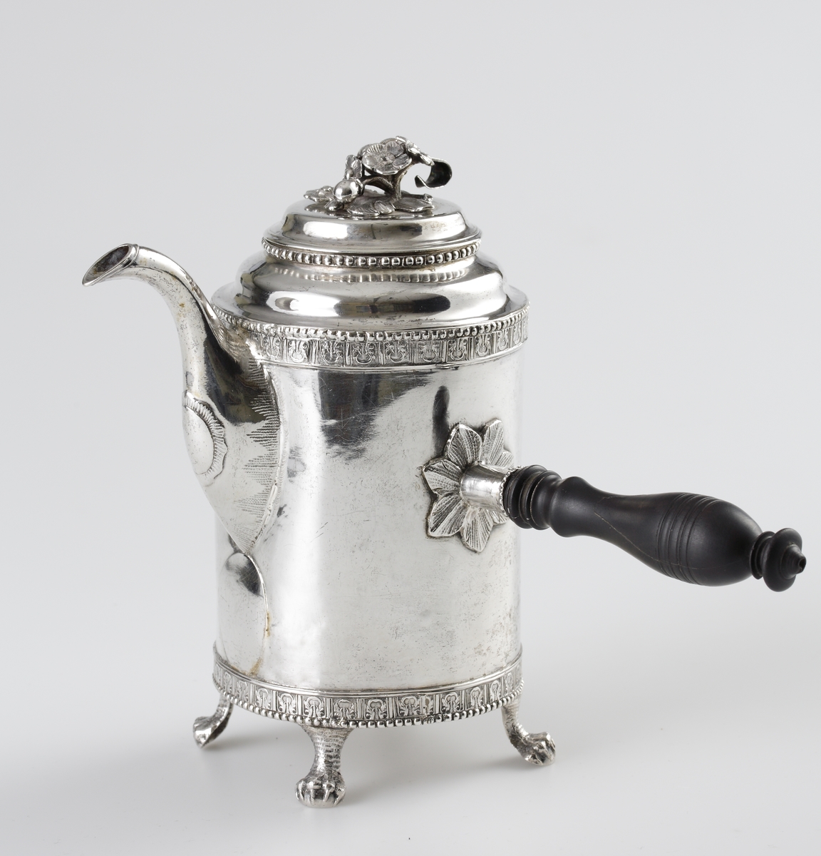 Kaffekanna i silver. 
Cylindrisk modell på fyra tassfötter. Pärlstavsdekor upptill och nedtill, innanför denna en ciselerad palmettbård. Välvt lock med graverad bård och knopp i form av blombukett. Kort, rakt utstående holk för skaftet. Vid holkens mynning dekor av pärlbård, vid dess inre kortsida omgiven av en pålödd och ciselerad blomma. 
Försedd med svarvat, svärtat trähandtag, fäst med ett bladformat böjt silverstift (saknas).
Stämplar på undersidan av kannan.

Inskrivet i huvudbok 1952.
