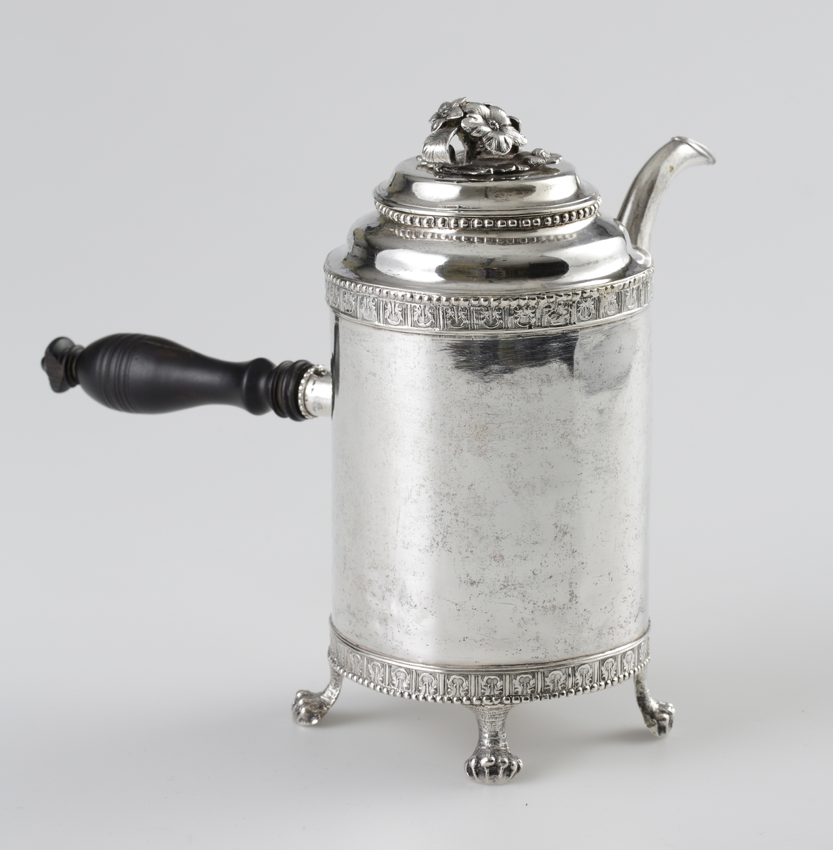 Kaffekanna i silver. 
Cylindrisk modell på fyra tassfötter. Pärlstavsdekor upptill och nedtill, innanför denna en ciselerad palmettbård. Välvt lock med graverad bård och knopp i form av blombukett. Kort, rakt utstående holk för skaftet. Vid holkens mynning dekor av pärlbård, vid dess inre kortsida omgiven av en pålödd och ciselerad blomma. 
Försedd med svarvat, svärtat trähandtag, fäst med ett bladformat böjt silverstift (saknas).
Stämplar på undersidan av kannan.

Inskrivet i huvudbok 1952.