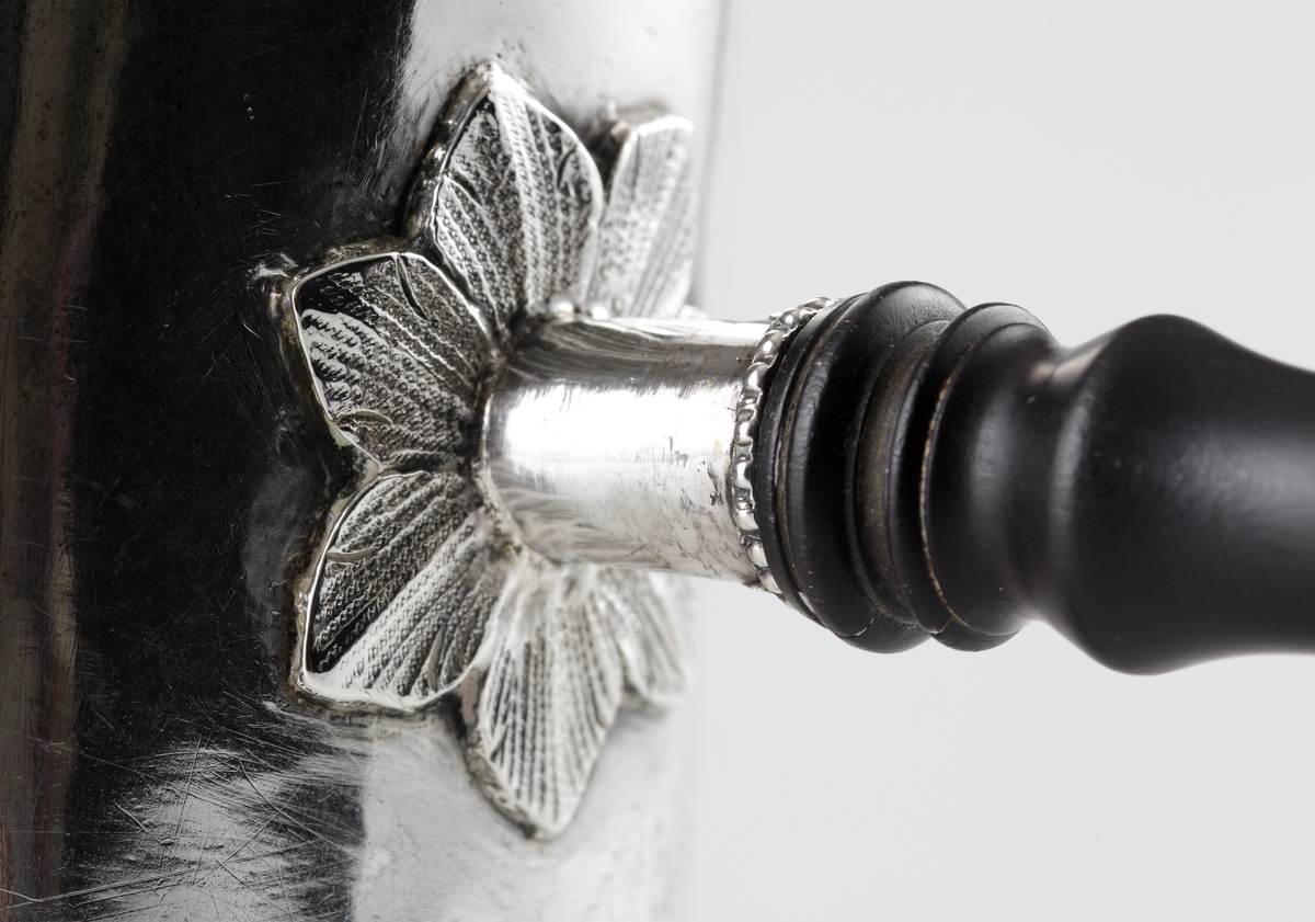Kaffekanna i silver. 
Cylindrisk modell på fyra tassfötter. Pärlstavsdekor upptill och nedtill, innanför denna en ciselerad palmettbård. Välvt lock med graverad bård och knopp i form av blombukett. Kort, rakt utstående holk för skaftet. Vid holkens mynning dekor av pärlbård, vid dess inre kortsida omgiven av en pålödd och ciselerad blomma. 
Försedd med svarvat, svärtat trähandtag, fäst med ett bladformat böjt silverstift (saknas).
Stämplar på undersidan av kannan.

Inskrivet i huvudbok 1952.