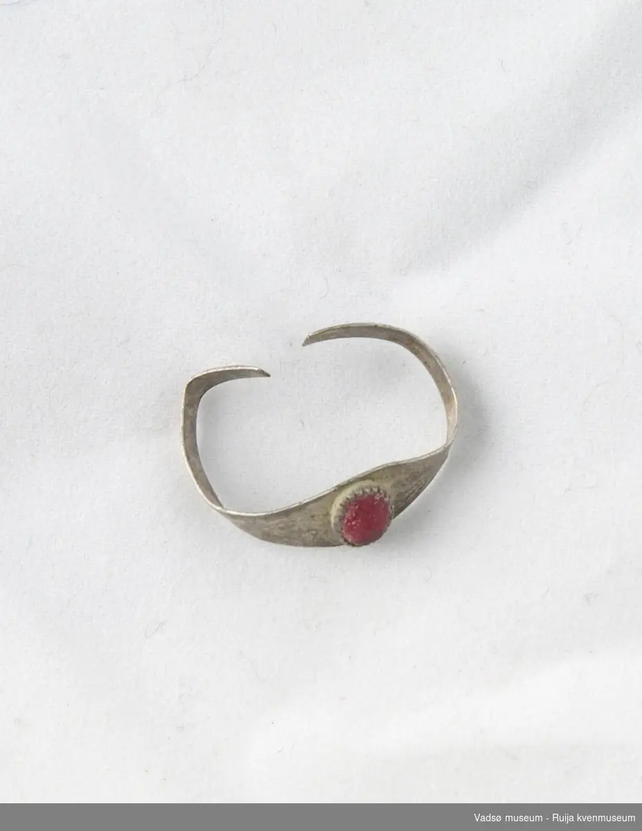 Sølvring som er dekorert med en oval, rød smykkestein av glass. Steinen har avrundete kanter, men viser tegn til opprinnelig fasettering. Sisellert prikke- og strekdekor på ringen.