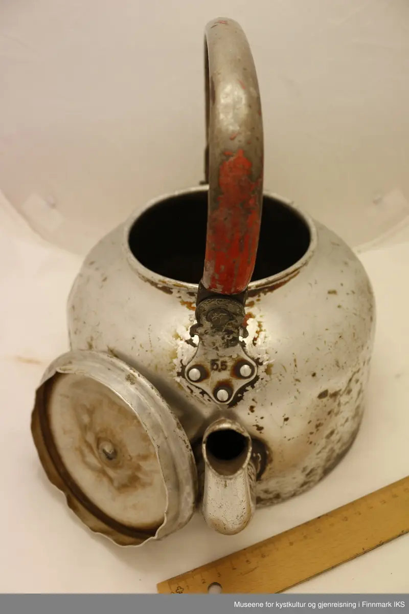 En stor kanne i aluminium med tut, håndtak og lokk. Håndtaket er rødmalt, men mye av malingen har flasset av. Kannen er produsert av Høyang.