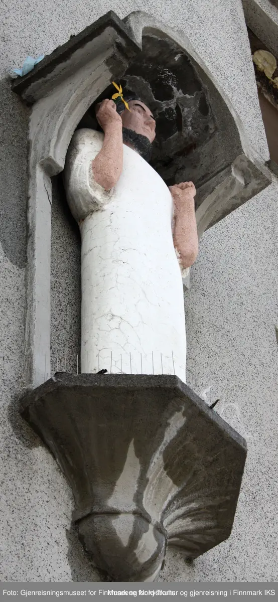 Figuren fremstiller en mannlig helgen med mørkt hår og lys drakt som holder en blomst, sannsynlig en lilje, i sin høyre hånd. Det finnes spor ved venstre hånd etter ett attributt til.
