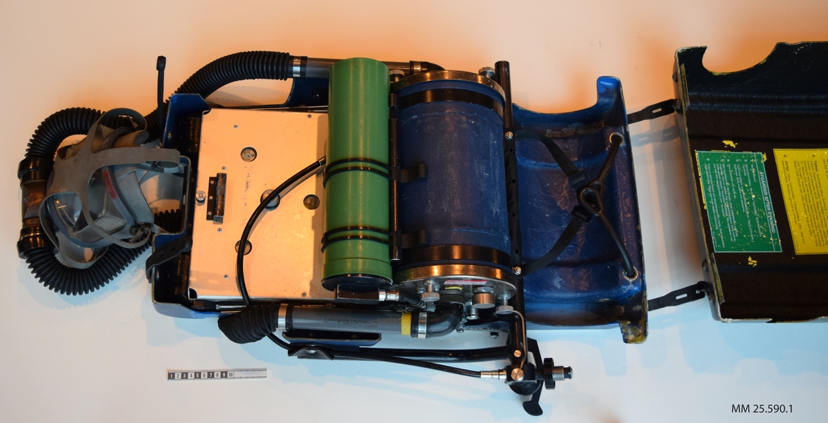 Dykapparat med andningsmask. Apparaten är fastsatt inom ett plasthölje vars form påminner om en resväska. Plasthäöjet öppnas opch stängs med hjälp av gummistroppar med hål som träs över skruvar/nitar. Inuti höljet finns två flaskor, en blå och en grön (doserflaska och blandgasflaska). Fäst i den blå flaskan löper två svarta gummislangar som går till andningsmasken. Masken är av grått gummi och täcker bärarens hela ansikte. Masken träs vöer ansiktet och runt bakhuvudet på bäraren sitter fyra gummidelar som förenas i en platta av gummi.