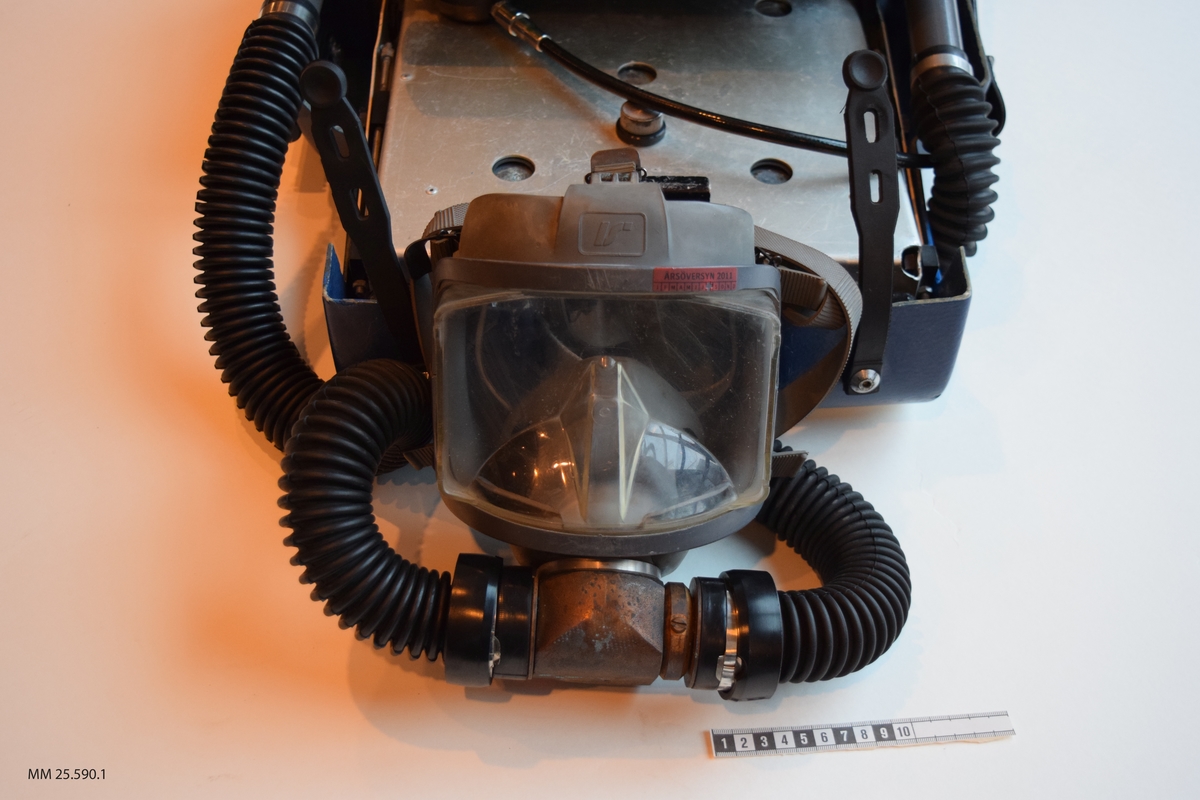 Dykapparat med andningsmask. Apparaten är fastsatt inom ett plasthölje vars form påminner om en resväska. Plasthäöjet öppnas opch stängs med hjälp av gummistroppar med hål som träs över skruvar/nitar. Inuti höljet finns två flaskor, en blå och en grön (doserflaska och blandgasflaska). Fäst i den blå flaskan löper två svarta gummislangar som går till andningsmasken. Masken är av grått gummi och täcker bärarens hela ansikte. Masken träs vöer ansiktet och runt bakhuvudet på bäraren sitter fyra gummidelar som förenas i en platta av gummi.