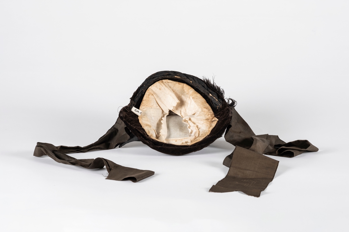 Rund hatt av plast trukket med fløyel. Hatten er pyntet med blondebånd, perler og fjær. Pullen er drapert med fløyel og ripsvevet bomull som ender i en sløyfe. Hatten har hakebånd. Svettebåndet er plastbelagt.