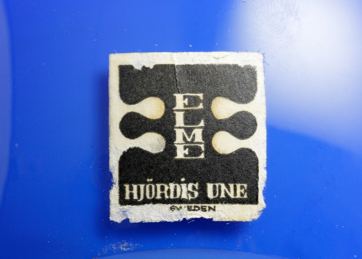 Blå skål i en rund och mjuk design. 
Etikett: Vit med ett stort svart dubbel-E med texten "Elme" och "Hjördis Une" i vitt.