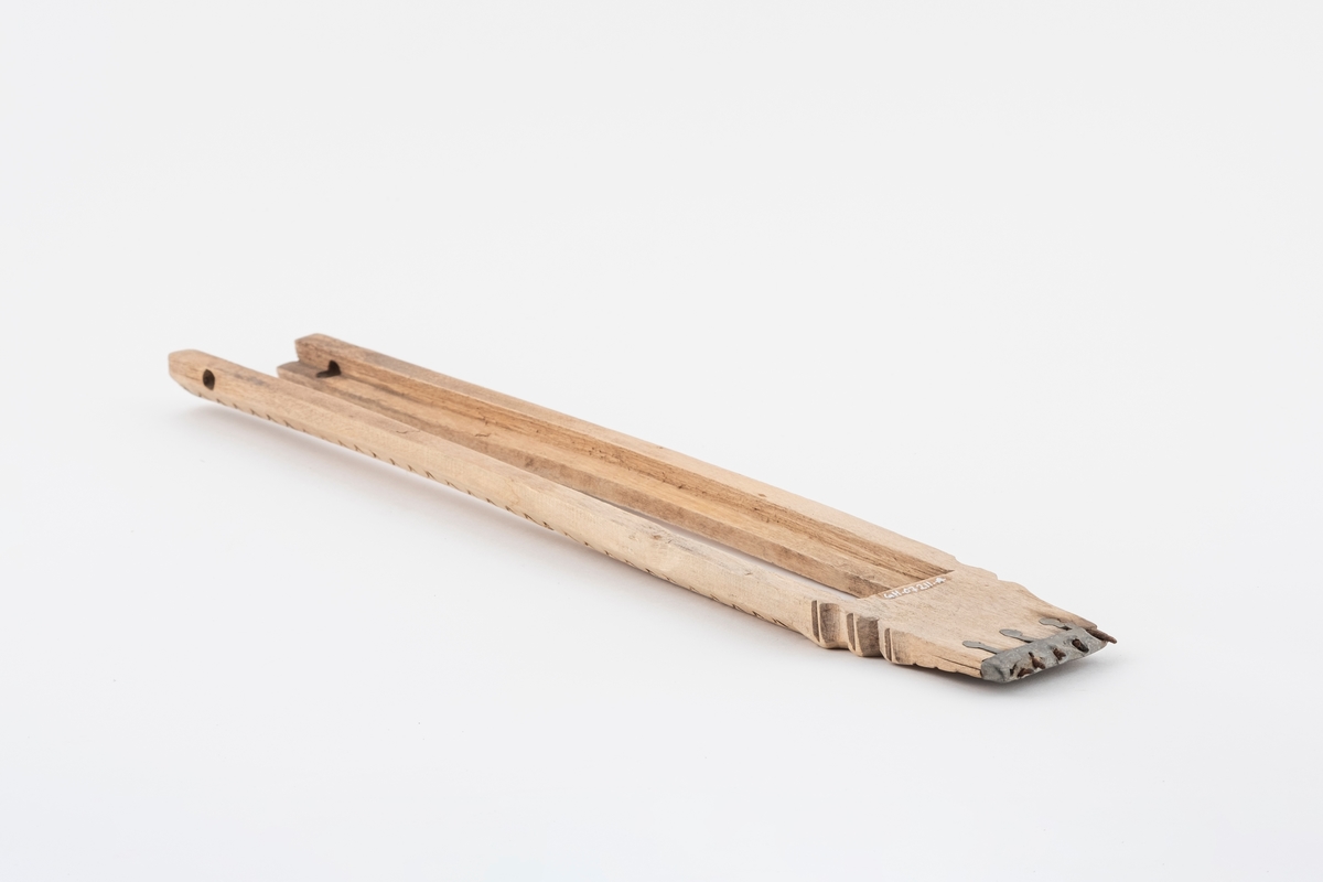 Del av vevspenne av tre, ytterdelen. Vevspenna har utskåret bølgeformet dekor, samt utskjæring i den ene enden. Det er et beslag av metall i den ene enden med pigger.