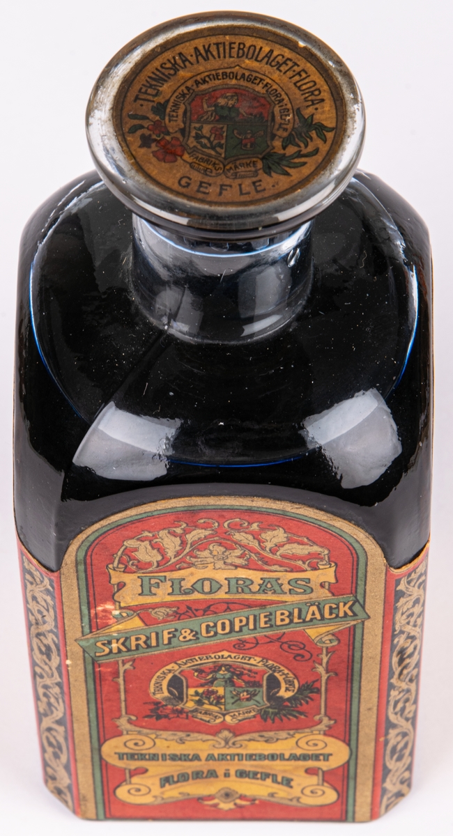 Bläckflaska med innehåll. Av glas. Oöppnad. Etiketter på samtliga sidor, varav framsidans i rött och med texten "FLORAS SKRIF & COPIEBLECK - TEKNISKA AKTIEBOLAGET FLORA I GEFLE", m.m.
Kvadratisk genomskärning med fasade hörn. Rundas upp till en smal rund hals med horisontell krage överst. Förseglad med pappetikett över korken. Etiketter på samtliga sidor, varav framsidans i rött med detaljer i flera färger. Sidoetiketterna har längre beskrivningar av bläcket på svenska resp. franska. På baksidan räknas prisbelöningar upp.