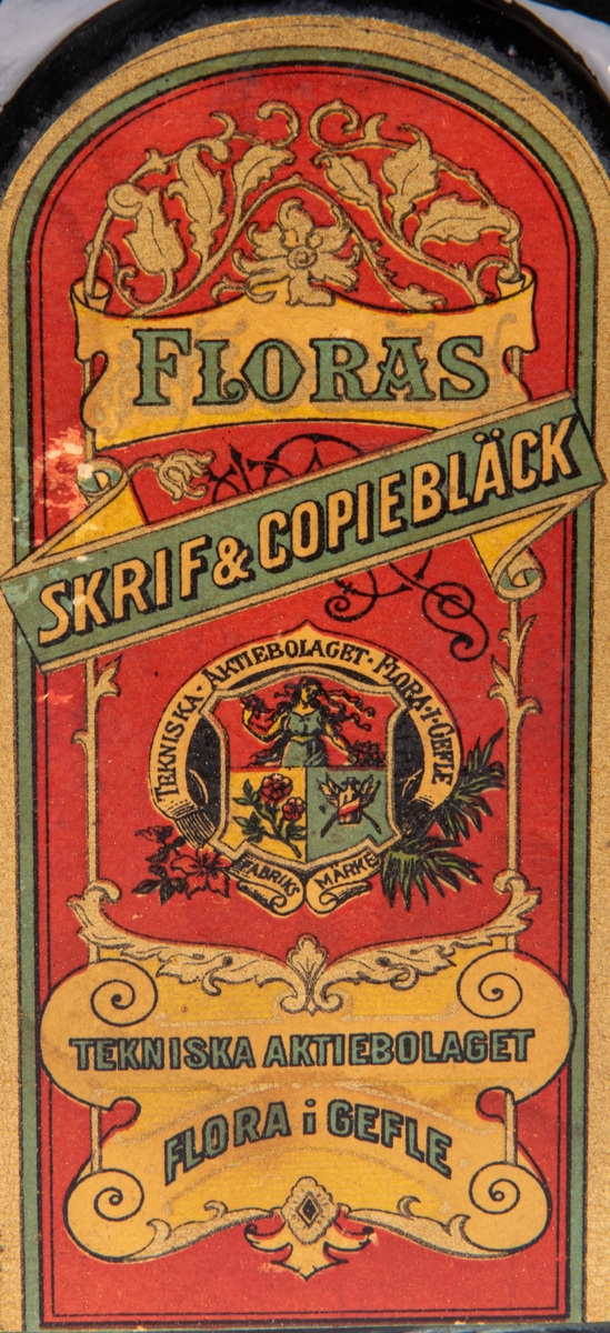 Bläckflaska med innehåll. Av glas. Oöppnad. Etiketter på samtliga sidor, varav framsidans i rött och med texten "FLORAS SKRIF & COPIEBLECK - TEKNISKA AKTIEBOLAGET FLORA I GEFLE", m.m.
Kvadratisk genomskärning med fasade hörn. Rundas upp till en smal rund hals med horisontell krage överst. Förseglad med pappetikett över korken. Etiketter på samtliga sidor, varav framsidans i rött med detaljer i flera färger. Sidoetiketterna har längre beskrivningar av bläcket på svenska resp. franska. På baksidan räknas prisbelöningar upp.