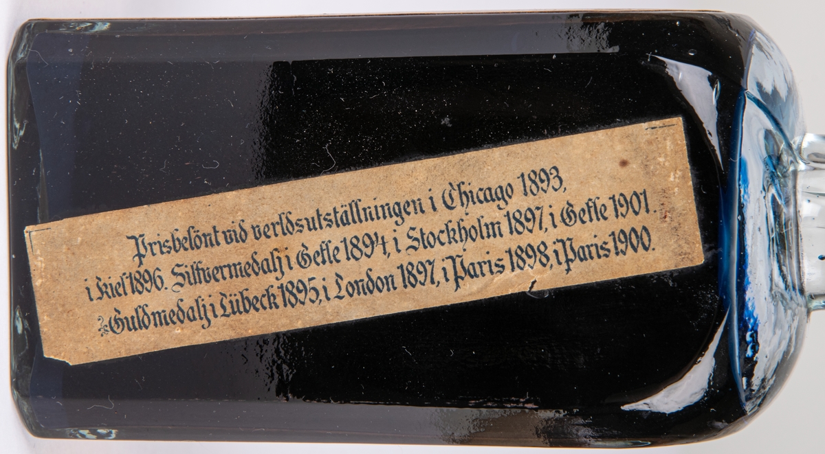 Bläckflaska med innehåll. Av glas. Oöppnad. Etiketter på samtliga sidor, varav framsidans i rött och med texten "FLORAS SKRIF & COPIEBLECK - TEKNISKA AKTIEBOLAGET FLORA I GEFLE", m.m.
Kvadratisk genomskärning med fasade hörn. Rundas upp till en smal rund hals med horisontell krage överst. Förseglad med pappetikett över korken. Etiketter på samtliga sidor, varav framsidans i rött med detaljer i flera färger. Sidoetiketterna har längre beskrivningar av bläcket på svenska resp. franska. På baksidan räknas prisbelöningar upp.