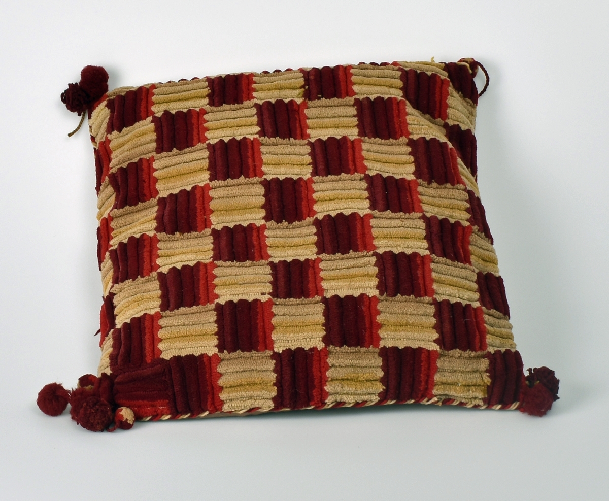 Sofapute. Forsiden har dekorative firkanter i flossteknikk. Kantene har påsydd snurret snor og i hjørnene er det dusker.  Baksiden har bomullsstoff med innvevd blomstermønster.