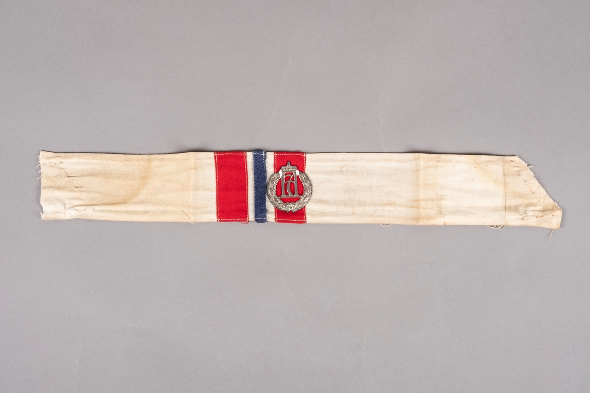 Et armbind i hvit bomull med påsydd rødt og blått tekstillapper. I midten er det påsydd Haakon 7. merke. Det er stempler på armbindet som er noe falmet.