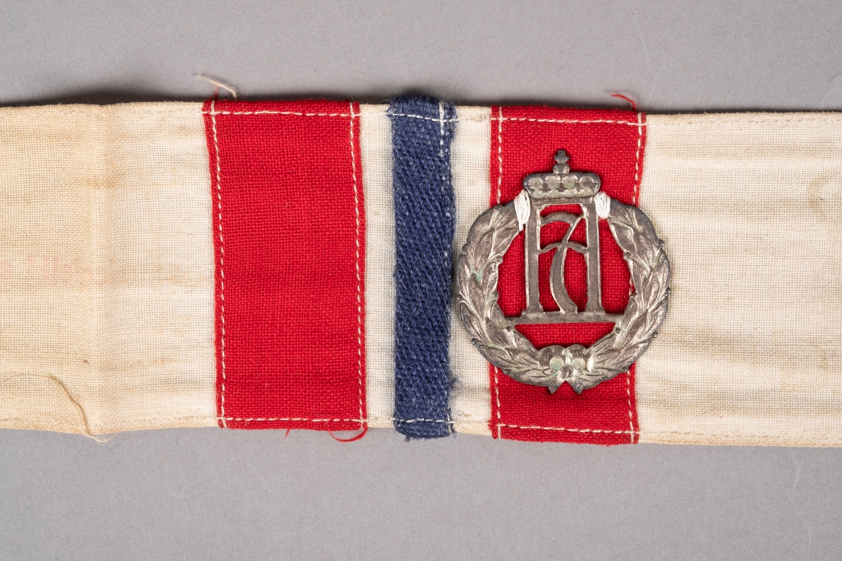 Et armbind i hvit bomull med påsydd rødt og blått tekstillapper. I midten er det påsydd Haakon 7. merke. Det er stempler på armbindet som er noe falmet.