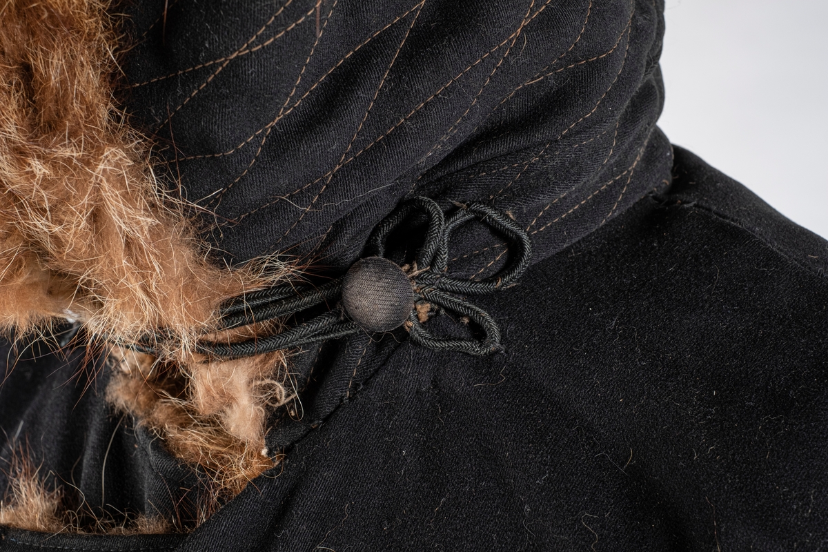 Frakk av bomull med pelsforing. Den har en bred krage. Kragen og mansjettene er kledd med pels. Frakken kneppes i halsen med knapp og hempe på hver side. Ved nedre kant er det brodert 3 røde kryss.