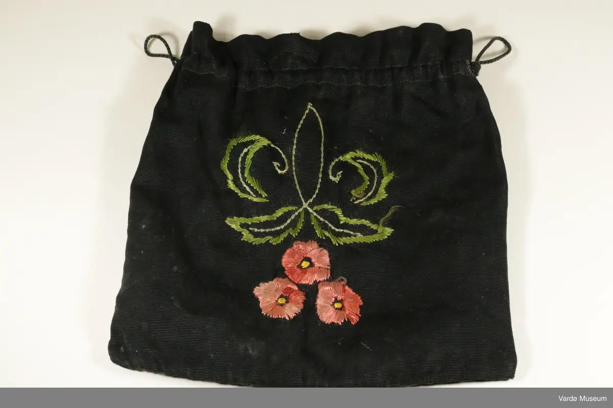 Sort pung i bomull. På den ene siden er det brodert inn tre blomster, med grønne blader rundt. På den andre siden er det ikke noe dekorasjon. Øverst er det snor på begge sidene, som man kan dra i for å lukke tøyposen igjen. 