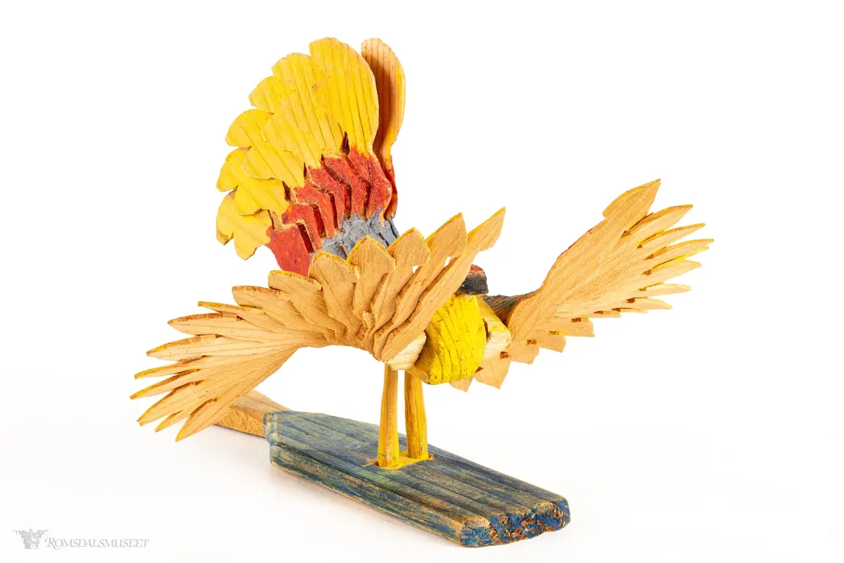 En sponfugl med livlige farger, med avtagbare vifteformede vinger, vifteformet hale og en avtagbar treplate med et lite håndtak. Kan være ment å være en liten vifte.