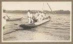 Bilden visar vita män i uniformer och civila kläder i en enkel segelbåt som har lagt till vid stranden för på- eller avstigning över en planka.