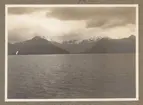 Bilden visar ett fjordlandskap med snötäckta berg och molnig himmel.