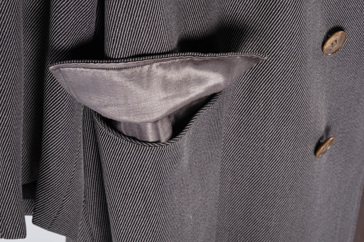 Rett frakk med slag og raglanerme. Foring av silke. Skjult knepping med fire knapper, kan også kneppe slaget under kragen. To lommer med klaff og to innerlommer. Splitt bak med knepping. Spensel nederst på ermene.